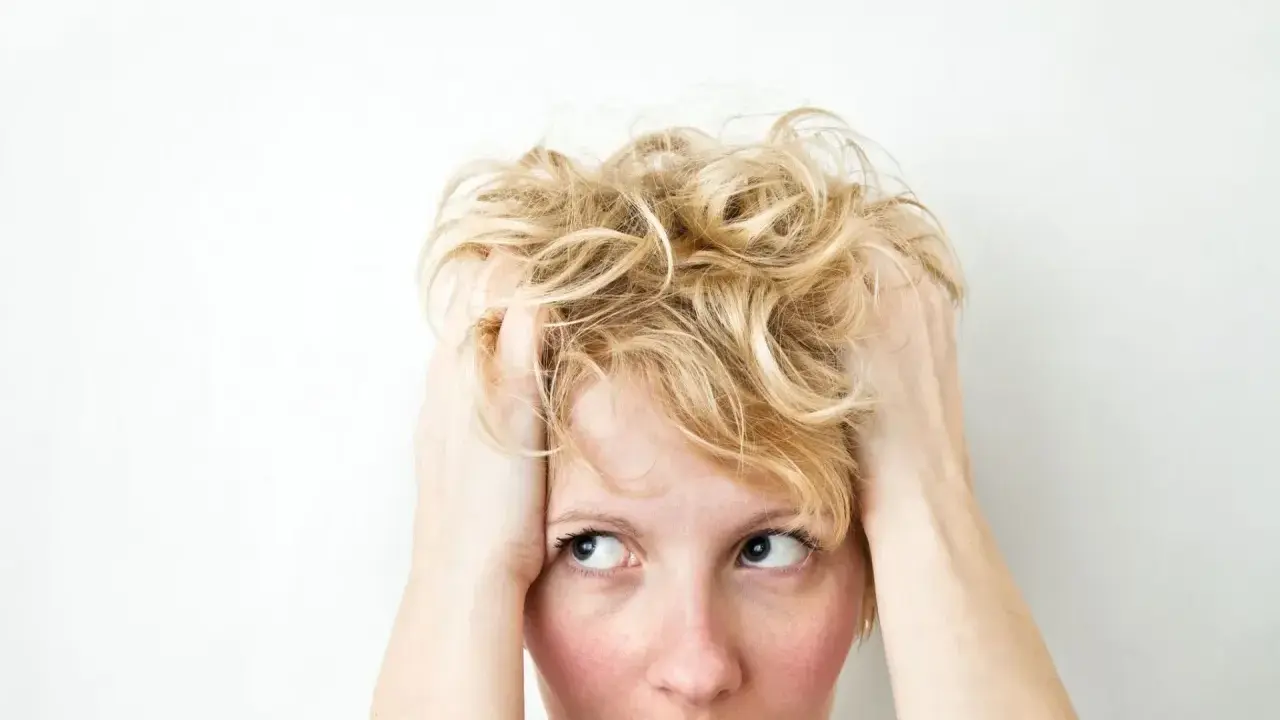 Kobieta z blond włosami wplata dłonie we włosy, jej wyraz twarzy sugeruje wahania nastroju i niepokój.