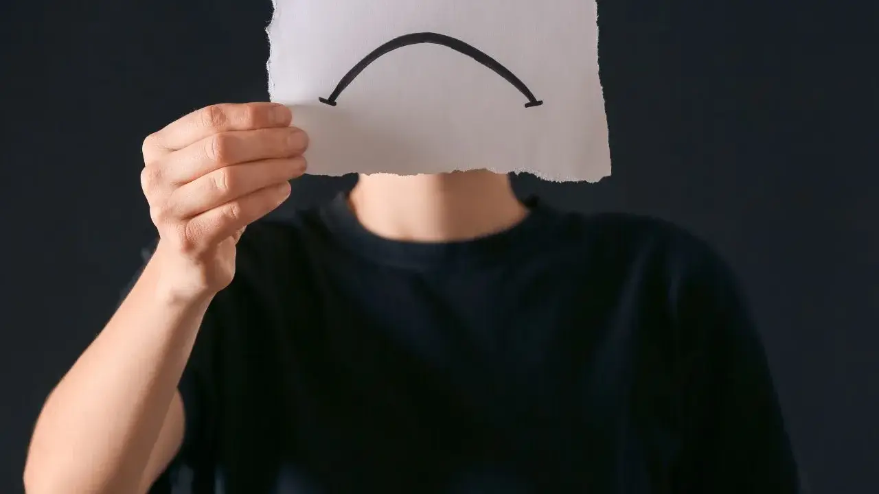 Osoba w czarnej bluzie trzyma kartkę z narysowaną smutną buźką. Czy to anhedonia, co to za uczucie?