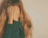 Kobieta z długimi blond włosami zakrywa twarz dłońmi na tle jasnej ściany.