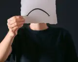Osoba w czarnej bluzie trzyma kartkę z narysowaną smutną buźką. Czy to anhedonia, co to za uczucie?