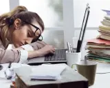 Kobieta zasnęła na laptopie, otoczona stertami dokumentów. To obraz pracoholizmu, który pochłania siły.
