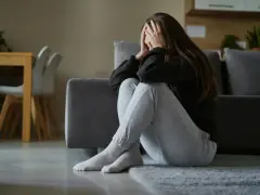 Kobieta siedzi na podłodze, zakrywając twarz dłońmi. Jej postawa sugeruje przygnębienie i może wskazywać na objawy somatyczne.