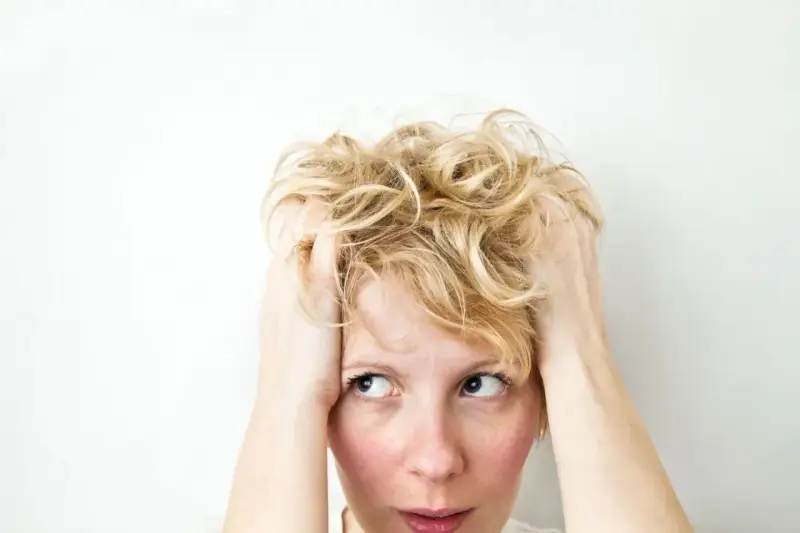 Kobieta z blond włosami wplata dłonie we włosy, jej wyraz twarzy sugeruje wahania nastroju i niepokój.